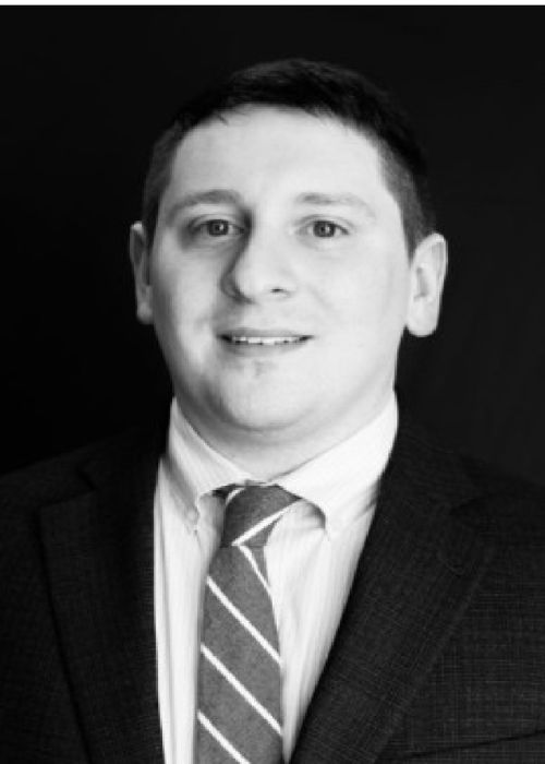 Black and white portrait of a man wearing a suit, striped tie, and collared shirt, looking at the camera and smiling slightly, with a dark background.