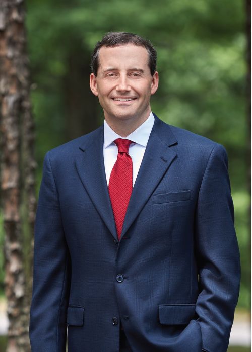 A man in a dark blue suit with a white shirt and red tie stands outdoors, smiling, with a background of green trees and blurred trunks.
