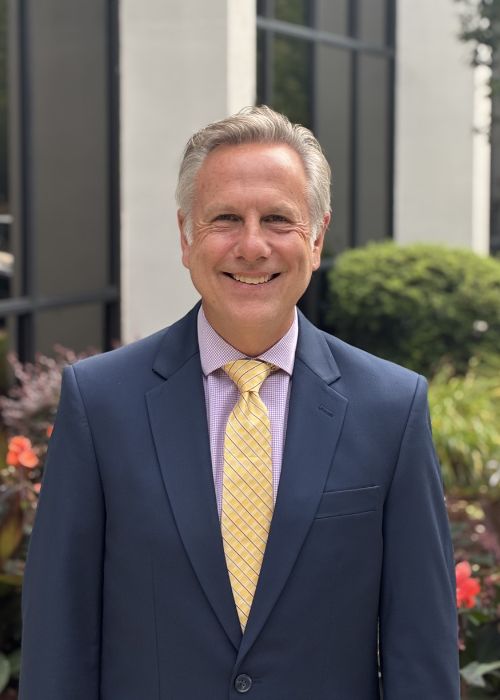 A smiling man in a blue suit, light purple shirt, and yellow striped tie stands outdoors in front of a building with glass windows and greenery.